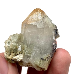 May include: A clear quartz crystal with a brown-tipped point. The crystal is mounted on a matrix of light green and beige minerals. The specimen is held in a hand against a white background.