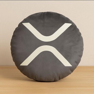 May include: A circular, grey cushion featuring the white XRP cryptocurrency symbol. The cushion is made of a soft material and rests on a wooden surface. The XRP symbol is a white, stylized design.