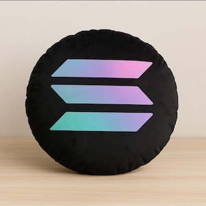 May include: Black round pillow featuring the Solana logo. The logo consists of three horizontal, overlapping, gradient-filled shapes in shades of pink, purple, and teal. The pillow is resting on a light-colored wooden surface.