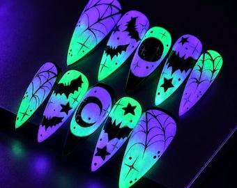May include: A set of ten stiletto-shaped press-on nails featuring a glow-in-the-dark finish. The designs include black spider webs, flying bats, crescent moons, and stars against a vibrant neon green and purple luminescent background.
