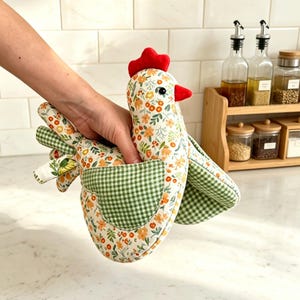 Può includere: Un guanto da forno a forma di gallina con una base in tessuto bianco caratterizzata da un motivo floreale di fiori arancioni e gialli e foglie verdi. Il guanto presenta un'ala e una coda a quadretti vichy verdi e bianchi, con cresta e becco rossi.