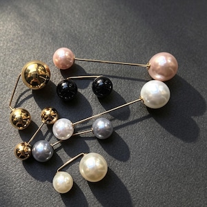 Könnte beinhalten: Sieben Broschen mit goldfarbenen Metallnadeln und kugelförmigen Akzenten in verschiedenen Farben, darunter Weiß, Rosa, Schwarz, Grau und Metallic-Gold. Der Text "Real photos of 7 pearl brooches" ist oben im Bild zu sehen.