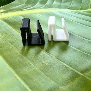 May include: Two small U-shaped clips, one in matte black and one in smooth white, featuring a rigid structure with parallel sides and a flat top. These functional components are designed with a distinct channel opening for securing items in place.