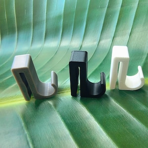 May include: Three small, U-shaped plastic hooks in gray, black, and white. Each hook features a flat top section, a vertical back panel, and a curved front hook designed for hanging items. The objects have a matte finish and a uniform, rigid structure.
