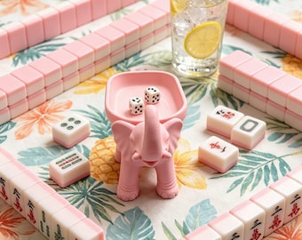 May include: A pink elephant-shaped dice tray holding two white dice, surrounded by rows of pink and white mahjong tiles. A clear glass filled with ice and lemon slices sits nearby on a table with a tropical floral and pineapple print pattern.