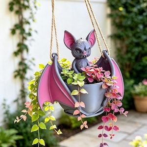 May include: A hanging planter shaped like a grey bat with large pink-lined ears and wings, holding a central grey pot. The bat has a textured grey body, a small pink nose, and two visible fangs. Trailing green and pink vines spill from the pot.