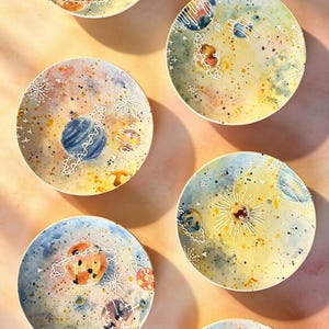 May include: A collection of six round decorative plates with a watercolor design of planets and stars. The plates feature a yellow, blue, orange, and purple color palette with white accents. The plates are arranged on a peach-colored surface.