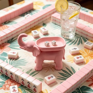 May include: A pink elephant-shaped tray with two white dice inside, surrounded by a set of pink and white Mahjong tiles on a floral tablecloth. A clear glass of water with a lemon slice sits nearby, completing the game setup on the table.