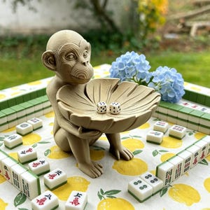 May include: A light brown monkey figurine holding a large, leaf-shaped bowl. Inside the bowl are two small white dice with black pips. The monkey has a textured, layered appearance and is positioned among several white and green mahjong tiles.