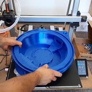 May include: A large, circular, bright blue 3D printed object with a hollow, bowl-like interior and a wide, flat rim. The object features internal structural dividers and a small circular opening on the edge, positioned on the print bed of a 3D printer.