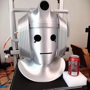 May include: A silver-colored Cyberman helmet replica featuring a smooth, metallic finish, prominent side ear-pieces, and cutouts for eyes and mouth. A standard 12-ounce soda can is placed next to the helmet for scale on a black printing platform.