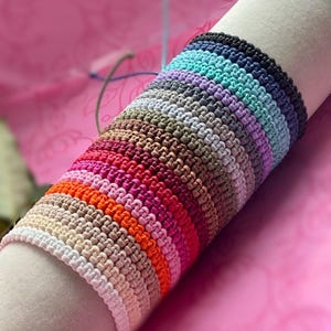 May include: A collection of colorful macrame bracelets in various shades, including white, beige, orange, pink, red, brown, purple, teal, and black. The bracelets are displayed on a white cylindrical form against a pink background.