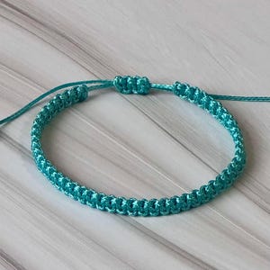 Könnte beinhalten: Ein türkisfarbenes Makramee-Armband mit verstellbarem Schiebeknotenverschluss. Das gewebte Design weist eine Reihe kleiner Knoten auf, die eine strukturierte Oberfläche bilden. Das Armband wird auf einer hellen Holzmaserung präsentiert.
