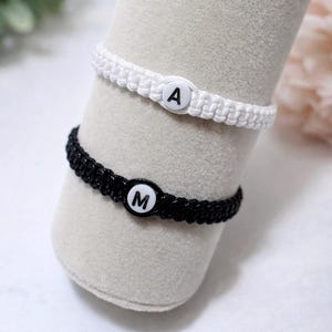 Könnte beinhalten: Zwei Makramee-Armbänder, eines weiß und eines schwarz, jeweils mit einer Buchstabenperle. Das weiße Armband hat ein "A" und das schwarze Armband ein "M". Die Armbänder werden auf einer zylindrischen, hellfarbigen Oberfläche präsentiert.