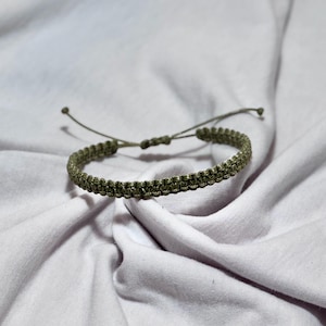 Puede incluir: Pulsera de macramé verde oliva. La pulsera está hecha de cordón tejido y presenta un cierre de nudo corredizo ajustable. La pulsera se muestra sobre un fondo blanco.