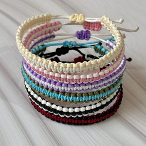 May include: A stack of handmade macrame bracelets in various colors, including cream, pink, purple, blue, and black. Each bracelet features a woven design with adjustable closures. The bracelets are stacked on a light-colored surface.
