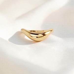 May include: A gold-colored ring with a smooth, curved design. The ring has a polished finish and a wave-like shape, reflecting light. The jewelry is set against a white background.