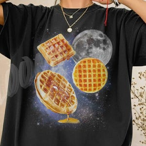 May include: Black t-shirt featuring a graphic of waffles, a moon, and a galaxy background. The waffles are depicted with syrup and a golden-brown color. The shirt has the text "FLOOMI co" on the lower right.