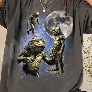 Può includere: T-shirt Comfort Color grigio scuro con una grafica di tre rane antropomorfe in una scena cosmica con una luna piena. La maglietta ha il testo "Comfort Color Shirt Pepper".
