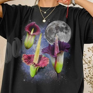 May include: Black t-shirt featuring a vibrant graphic of three stylized flowers with purple, pink, and green hues, set against a backdrop of a full moon and a starry night sky. The shirt has a relaxed fit.