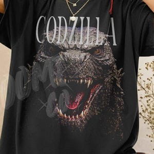 May include: Black t-shirt featuring a detailed graphic of Godzilla with the word "GODZILLA" in white block letters above the monster's head. The shirt has a vintage, slightly faded look.