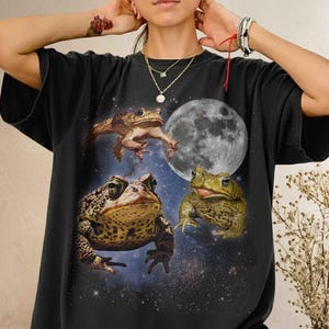 May include: A black t-shirt featuring a graphic print of three toads positioned against a backdrop of a full moon and a starry night sky. The design includes one toad leaping at the top, one large toad in the center, and one toad on the right.