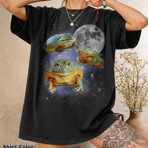 May include: Black t-shirt featuring a whimsical design of three colorful frogs against a starry night sky and a full moon. The shirt has the text "Shirt Color Black" at the bottom.