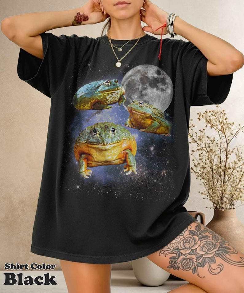 May include: Black t-shirt featuring a whimsical design of three colorful frogs against a starry night sky and a full moon. The shirt has the text "Shirt Color Black" at the bottom.