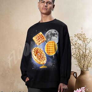 May include: Black sweatshirt featuring a whimsical design of waffles, a moon, and a starry background. The waffles appear to be floating in space, with one drizzled with syrup. The text "Sweatshirt Color BLACK" is at the bottom.