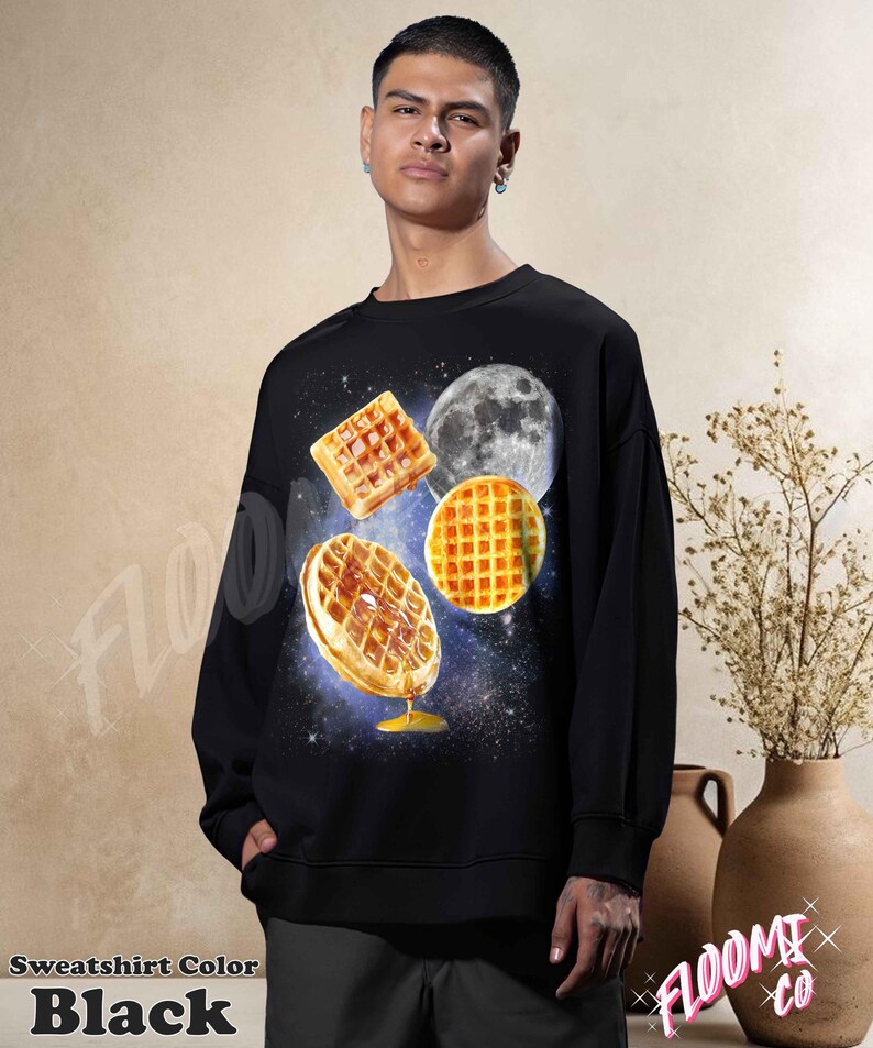 May include: Black sweatshirt featuring a whimsical design of waffles, a moon, and a starry background. The waffles appear to be floating in space, with one drizzled with syrup. The text "Sweatshirt Color BLACK" is at the bottom.