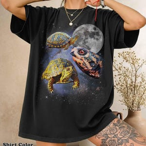 Puede incluir: Una camiseta negra de manga corta con un estampado gráfico de dos tortugas de caja y una cabeza de tortuga grande sobre un fondo de espacio estrellado con una luna llena. El diseño incluye caparazones amarillos y marrones.