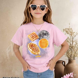 May include: Light pink t-shirt featuring a waffle and moon graphic. The shirt has a crew neck and short sleeves. The text "Shirt Color Light Pink" is at the bottom left of the image.