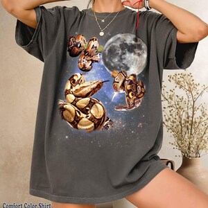 Può includere: Una t-shirt a maniche corte color grigio antracite con una stampa grafica di due serpenti dal motivo marrone e marrone chiaro avvolti attorno a una grande luna piena dettagliata su uno sfondo di spazio stellato blu scuro.