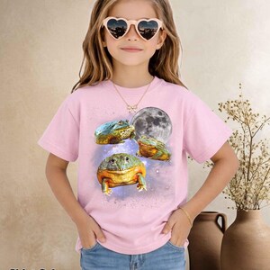 May include: Light pink t-shirt featuring a colorful frog design with a full moon graphic. The shirt has short sleeves and is worn by a young person. The text "Shirt Color Light Pink" is at the bottom of the image.