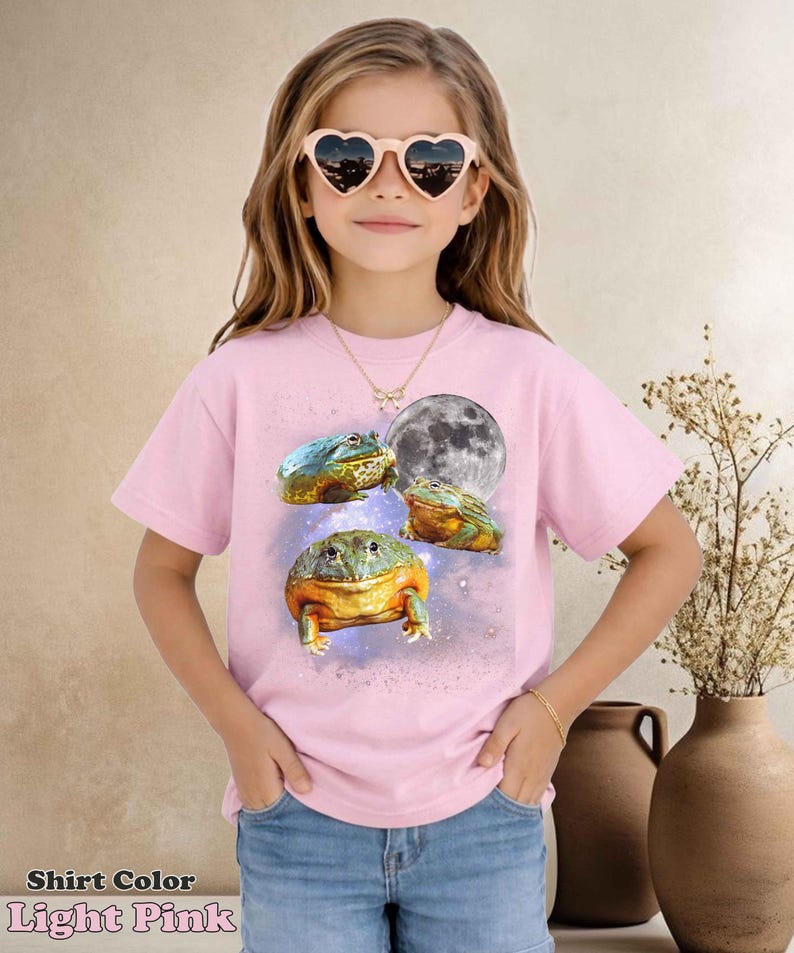 May include: Light pink t-shirt featuring a colorful frog design with a full moon graphic. The shirt has short sleeves and is worn by a young person. The text "Shirt Color Light Pink" is at the bottom of the image.