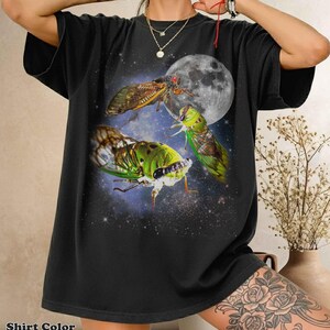 Peut inclure: Un t-shirt noir avec une impression graphique représentant trois cigales dans des tons de vert et de brun sur un fond d'espace sombre avec une grande lune détaillée. Les insectes présentent des motifs d'ailes complexes.