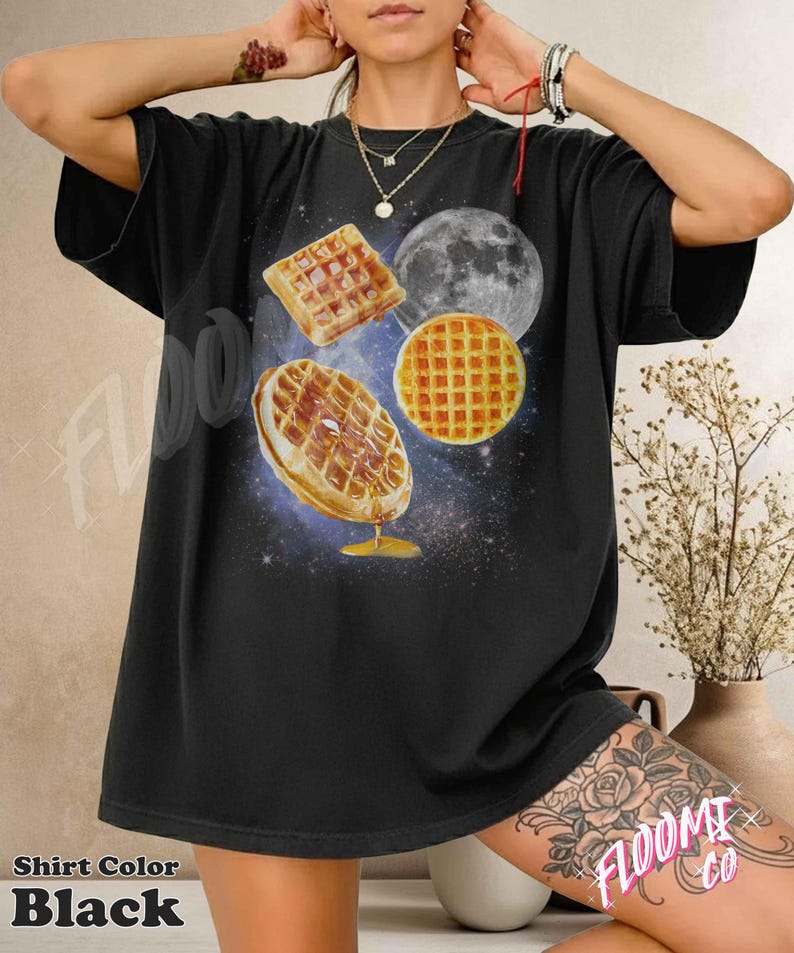 May include: Black t-shirt featuring a graphic of waffles, a moon, and a galaxy background. The waffles are depicted with syrup and a golden-brown color. The shirt has the text "FLOOMI co" on the lower right.
