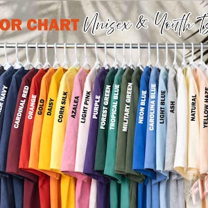 May include: A color chart of Unisex & Youth T-shirts in various colors, including black, navy, red, orange, yellow, pink, purple, green, blue, and white. Each shirt is labeled with its color name. The shirts are hanging on a metal rack.