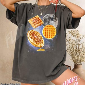 Può includere: Una t-shirt grigio antracite con una stampa grafica di tre waffle dorati e una luna piena su uno sfondo spaziale blu scuro stellato. Il design include sciroppo che cola da un waffle e sottili scintillii sparsi in tutta la scena.