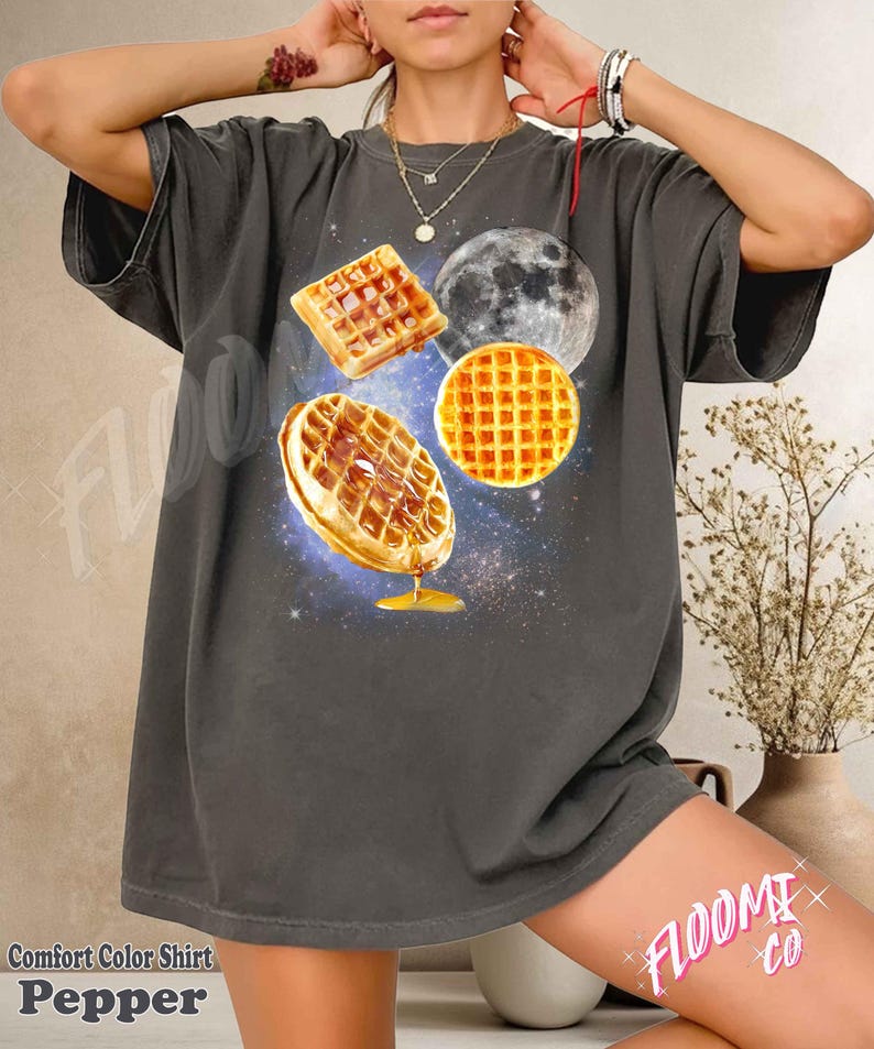 May include: Dark grey Comfort Color t-shirt with a graphic of waffles, a moon, and a galaxy background. The waffles are depicted with syrup dripping down. The text "FLOOMI CO" is visible on the shirt.