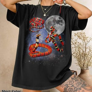Puede incluir: Una camiseta negra de manga corta con un estampado gráfico de tres serpientes rojas, amarillas y negras enroscadas sobre un fondo de luna llena y una galaxia estrellada. La prenda presenta un cuello redondo y un corte holgado y relajado.