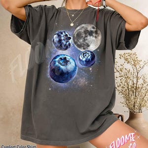 May include: A charcoal grey short-sleeve t-shirt featuring a graphic print of three blueberries and a full moon set against a starry cosmic background. The shirt has a crew neckline and a relaxed fit, with the text Floomi Co visible on the design.