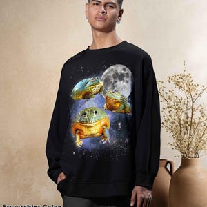 May include: Black sweatshirt featuring a whimsical design of three colorful frogs set against a starry galaxy background with a full moon. The text "Sweatshirt Color Black" is at the bottom of the image. A unique and eye-catching apparel item.