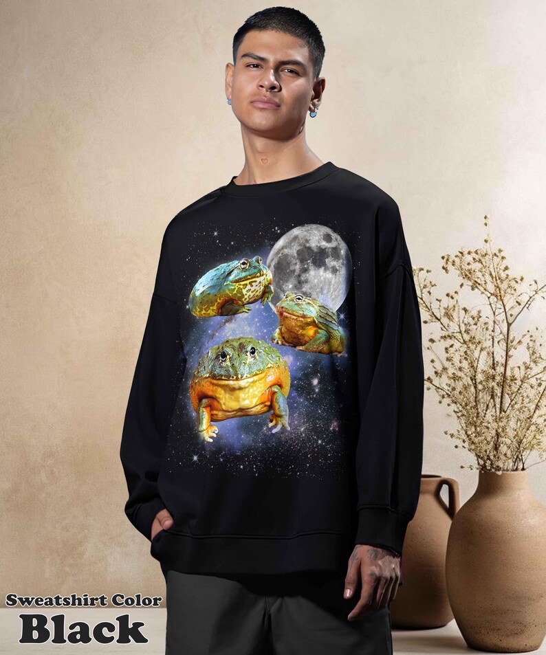 May include: Black sweatshirt featuring a whimsical design of three colorful frogs set against a starry galaxy background with a full moon. The text "Sweatshirt Color Black" is at the bottom of the image. A unique and eye-catching apparel item.
