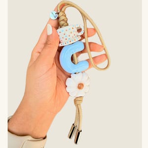 May include: A beige cord phone charm with a blue letter "C", a white daisy, and a small teacup charm. The charm has gold-colored metal accents and a small tag.
