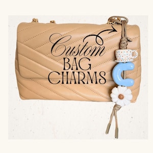 May include: A beige quilted handbag with a decorative bag charm. The charm includes a blue letter "C", a white daisy, and a small teacup. The text "Custom BAG CHARMS" is written on the bag.