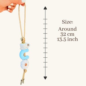 May include: A decorative charm with a light blue letter "C", a white flower, and a polka-dot design. The charm is suspended from a beige cord. The size is around 13.5 inches.
