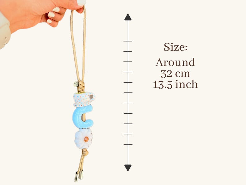 May include: A decorative charm with a light blue letter "C", a white flower, and a polka-dot design. The charm is suspended from a beige cord. The size is around 13.5 inches.