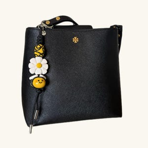 May include: Black handbag with a gold logo and a decorative charm. The charm features a yellow and white beaded design with a bee and flower motif. The bag has a top handle and a shoulder strap.