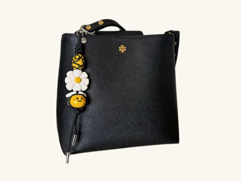 May include: Black handbag with a gold logo and a decorative charm. The charm features a yellow and white beaded design with a bee and flower motif. The bag has a top handle and a shoulder strap.